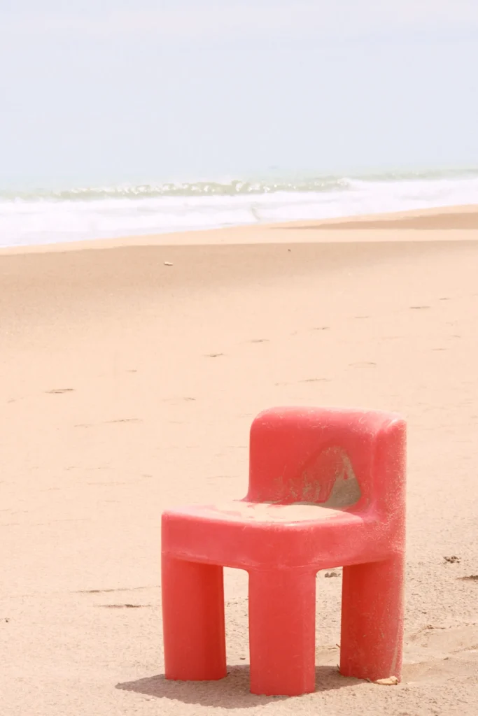 Minimalist destination lifestyle photo featuring a lone red plastic chair on an empty, soft-focus sandy beach, capturing a warm, cinematic travel mood in Spain or Southern Portugal