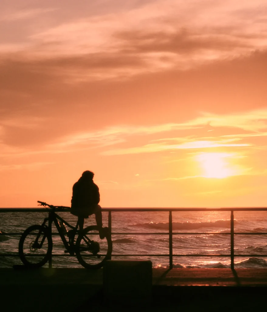 Moody and cinematic destination lifestyle photo of a beach promenade at sunset, showing human silhouettes and metal arches against an orange sky in Sitges, Spain.