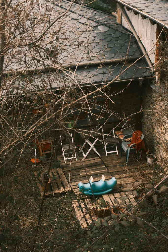 Moody and intimate rural lifestyle photography of a hidden outdoor patio area next to a stone cabin, with wooden pallets and scattered chairs, capturing a quiet destination feel in Andorra