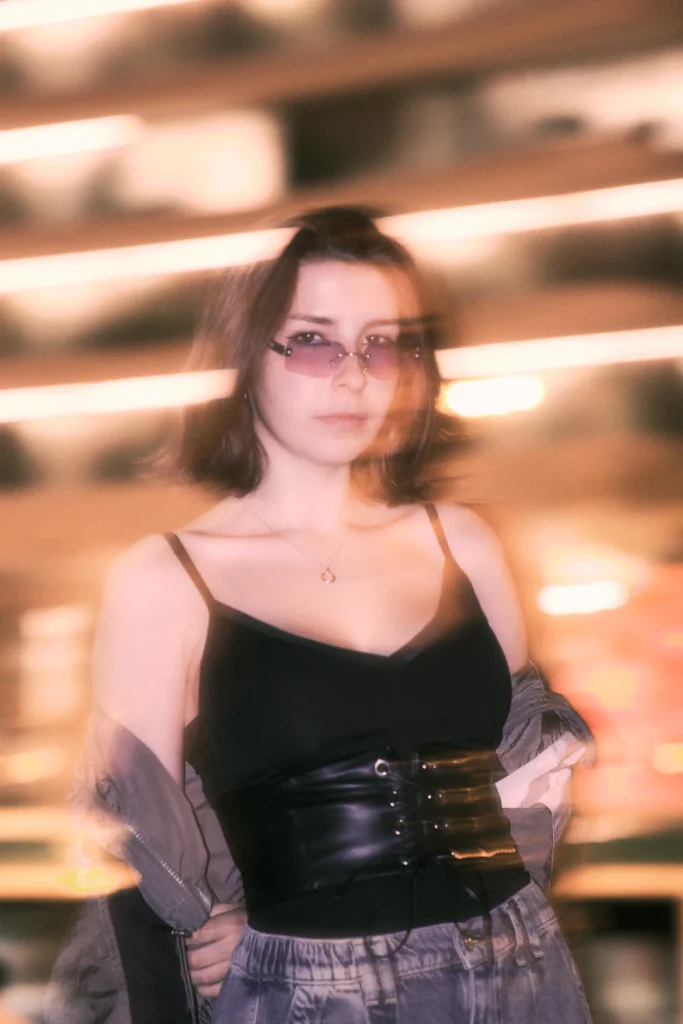 Fashion-focused night urban portrait of a woman in streetwear and small sunglasses, captured with intentional motion blur and neon glow effects.