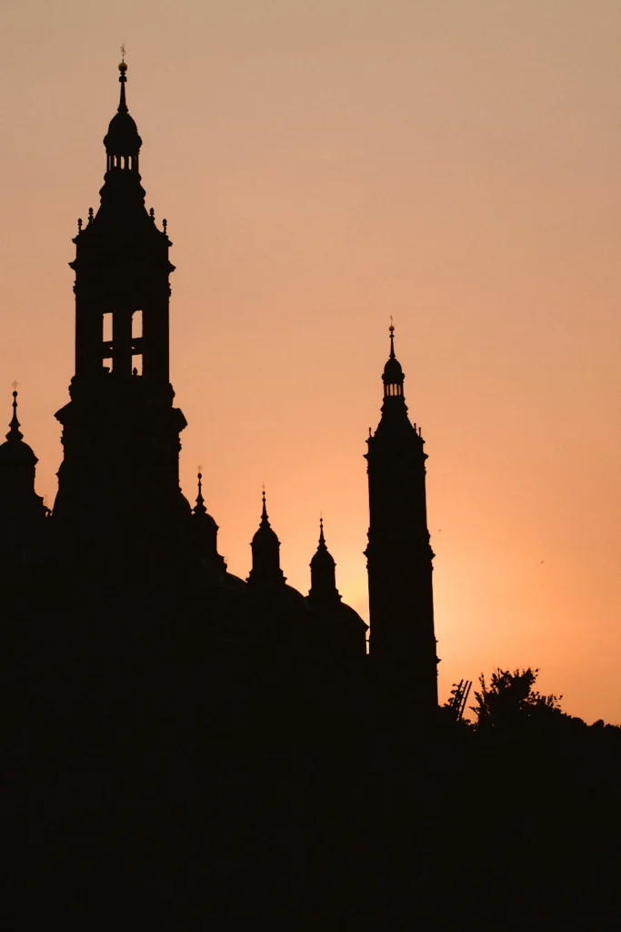 Dramatic silhouette of a major Spanish landmark (likely Basilica del Pilar) against a deep orange sunset sky, capturing cinematic destination photography in Zaragoza, Spain