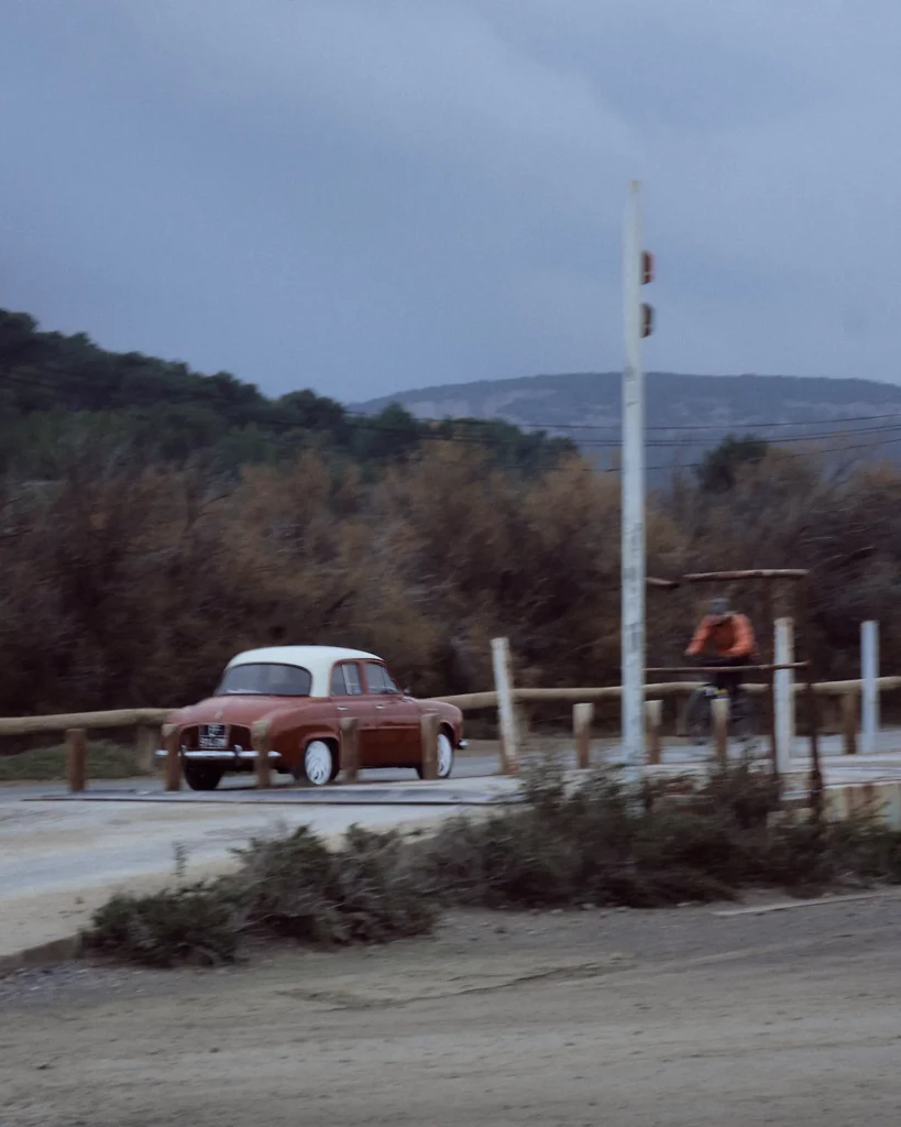 Scenic and moody cinematic travel photography of a vintage red and white car driving on a dirt road, framed by dry vegetation, emphasizing high-end travel destination content in France or Spain