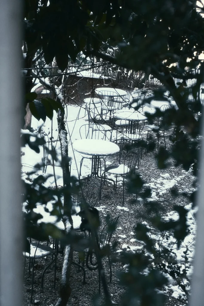Moody winter editorial photography of an empty snow-covered outdoor cafe seating area, framed by dark foliage, capturing a cold destination lifestyle mood in Switzerland or Eastern Europe