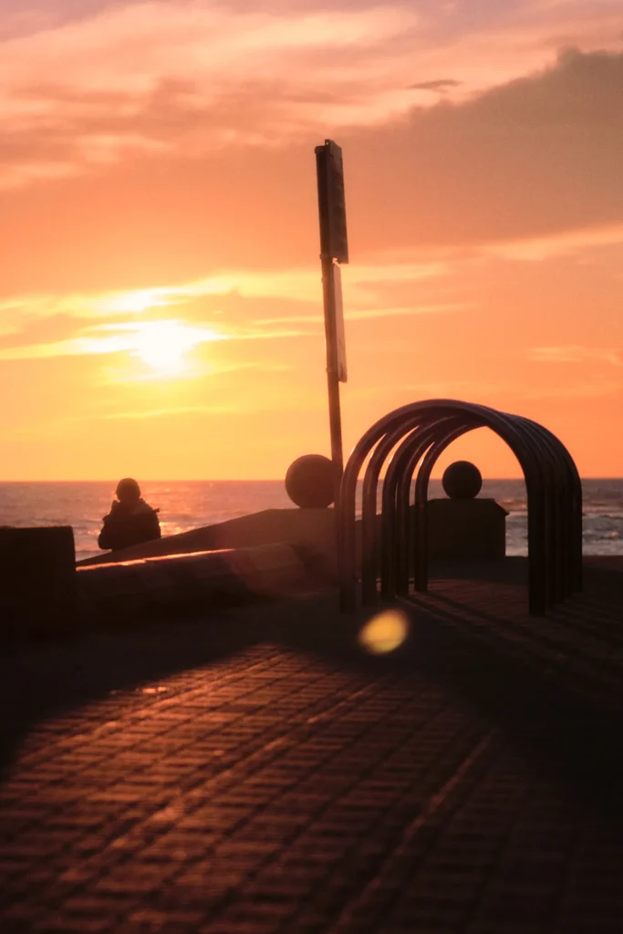 Golden hour cinematic photography of a person sitting on a bicycle silhouette, overlooking the ocean from a beach promenade railing at sunset in Sitges, Spain