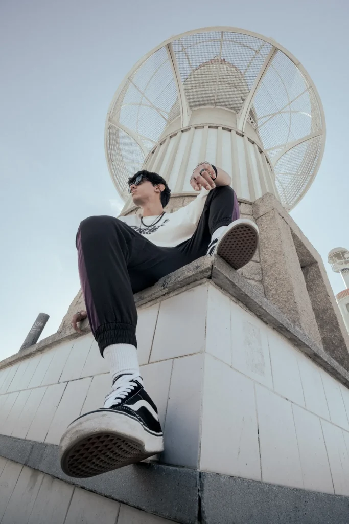 Low-angle urban fashion portrait of a young male model wearing streetwear, framed by a unique architectural skyline feature.