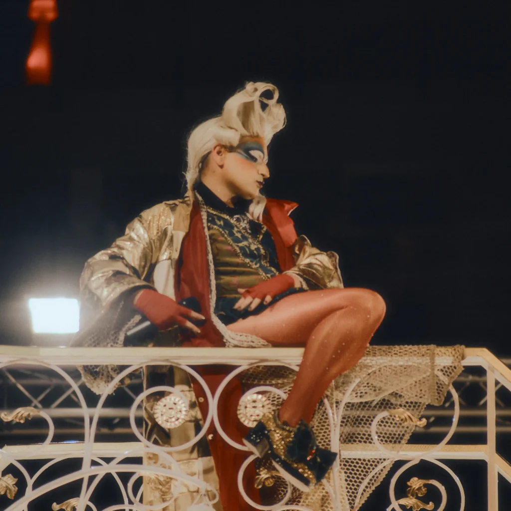 Cinematic high-end editorial lifestyle photography of a drag queen performer in gold and red costume on a stage, capturing vibrant nightlife and event content in Sitges, Spain.