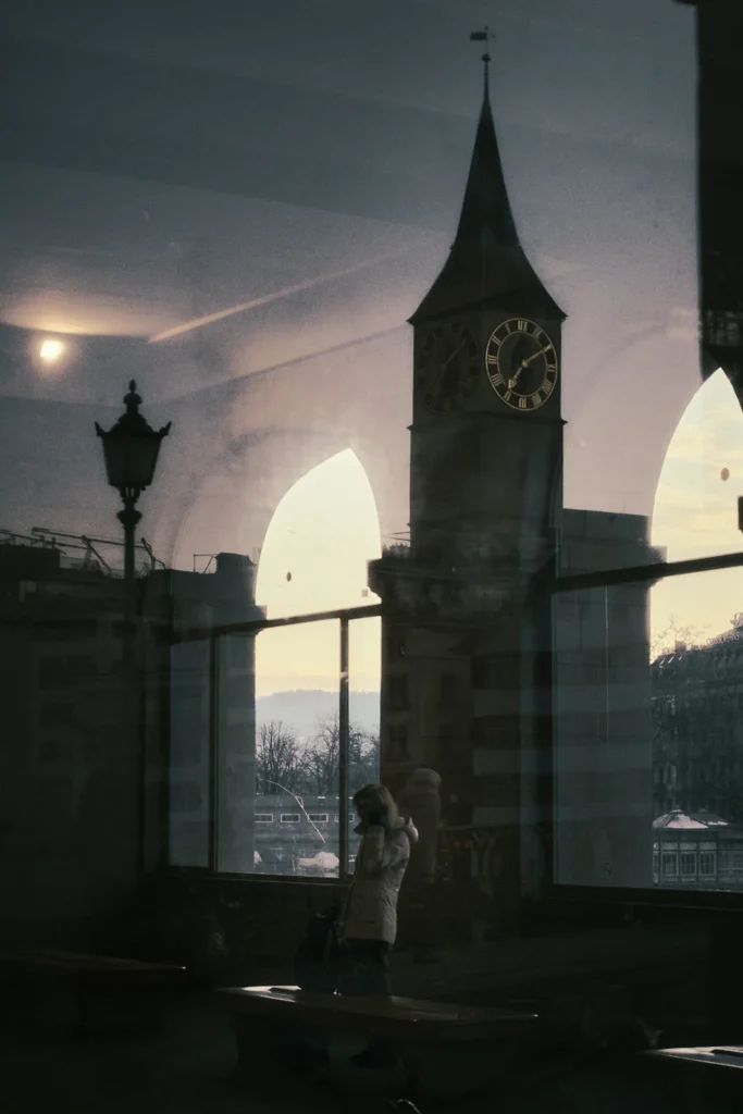 Moody and cinematic city portrait photography of a person standing by a window, featuring a reflection of the Zurich clock tower at sunset, emphasizing high-end lifestyle travel photography
